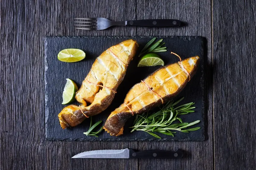 Two smoked Halibut steaks are served with lime wedges and rosemary on a black slate beside a fork and knife.