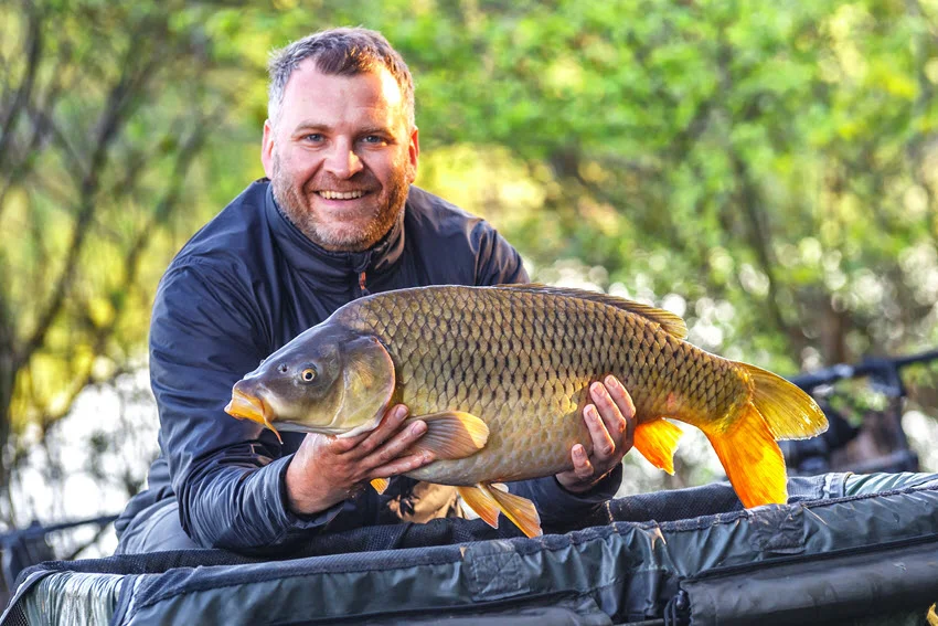 Angler holding a large Common Carp at a lake, proudly showing his trophy after a successful catch.