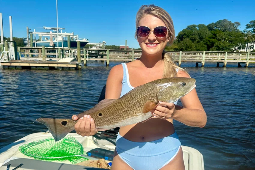Angler holding a Red Drum on a nearshore boat with docks and calm coastal water in the background.