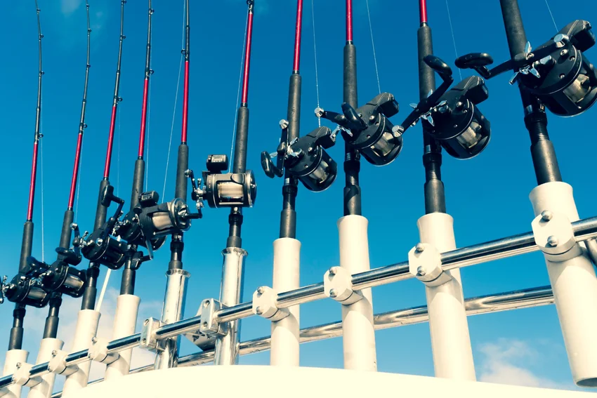 Conventional reels mounted on fishing rods stand in white holders along a boat rail, with their spools and handles visible against blue sky.