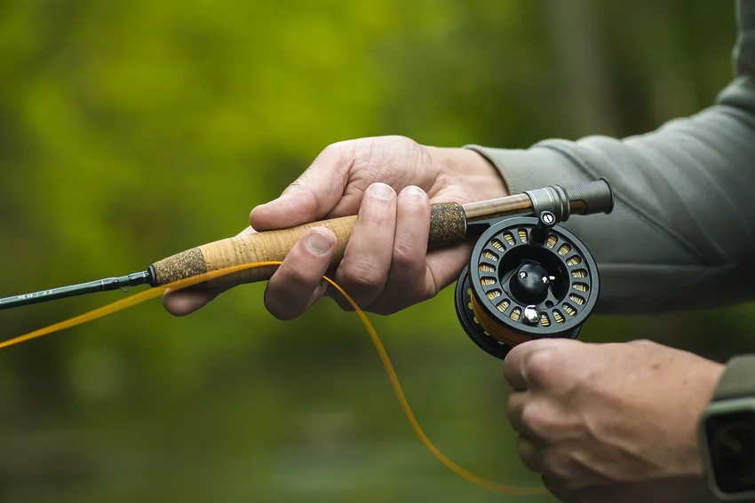 An angler holds a cork-handled fly rod with a round fly reel beneath it, while yellow line hangs loosely between the hands.