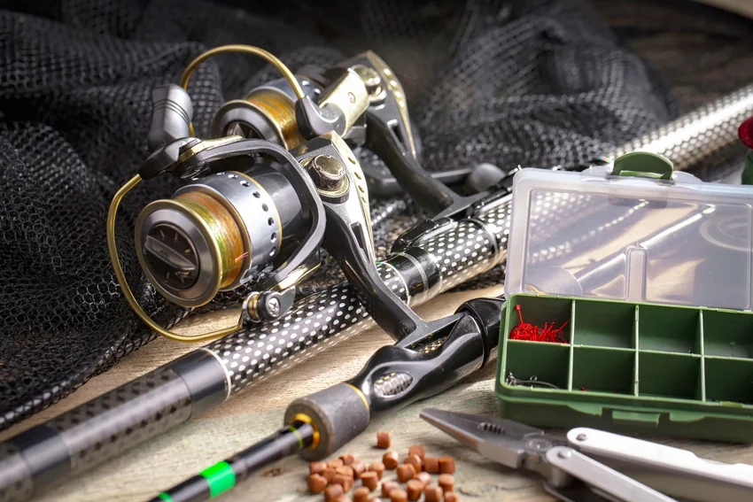 Two spinning reels mounted on fishing rods rest beside a landing net, an open tackle box, scattered bait pellets, and fishing pliers.