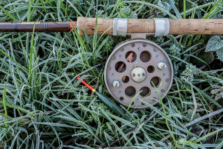 A vintage centerpin reel with two handles is mounted beneath a cork-handled rod, resting beside a red-tipped float on frost-covered grass.