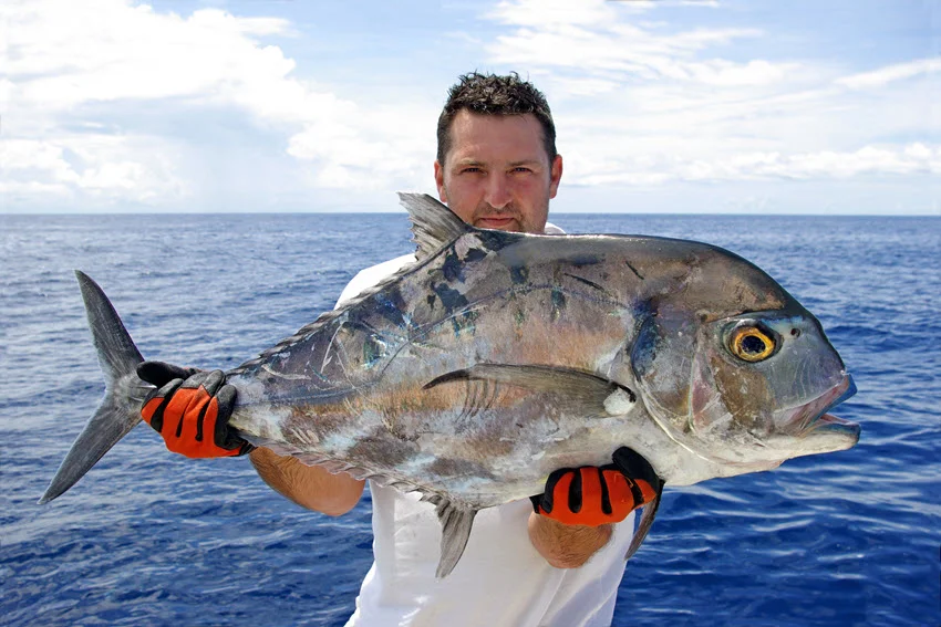 Angler holding a large Giant Trevally on an offshore boat with open blue water behind him.