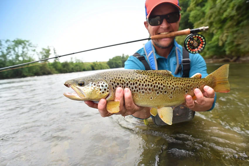 Fly angler holding a Brown Trout in a river, with the rod and reel visible behind the catch.