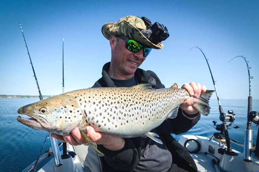Close view of an angler holding a Lake Trout on a boat with trolling rods set behind him on calm open water.