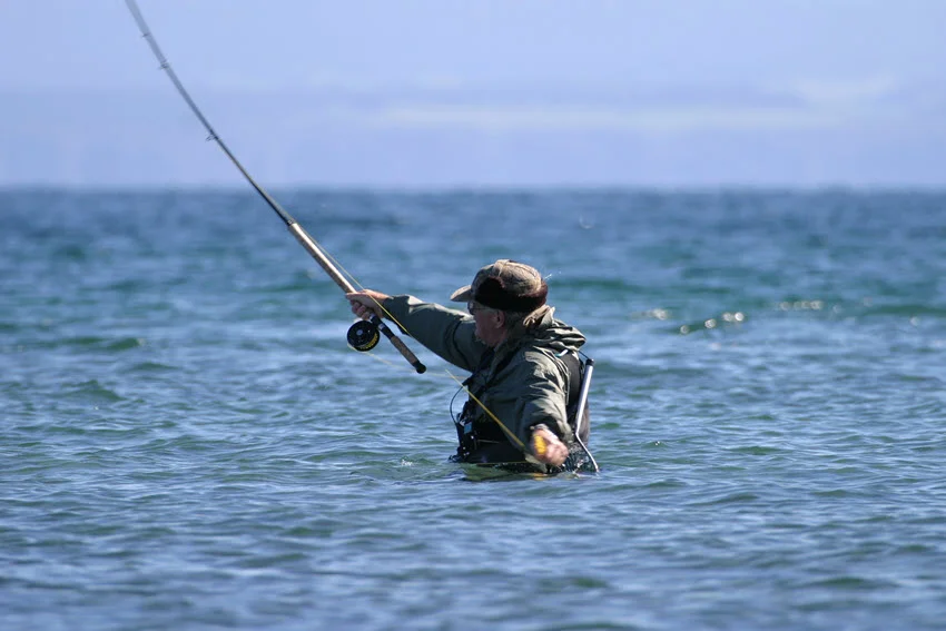 Close-up shot of a wade angler casting a rod while standing waist-deep in the sea, with waves gently surrounding him.