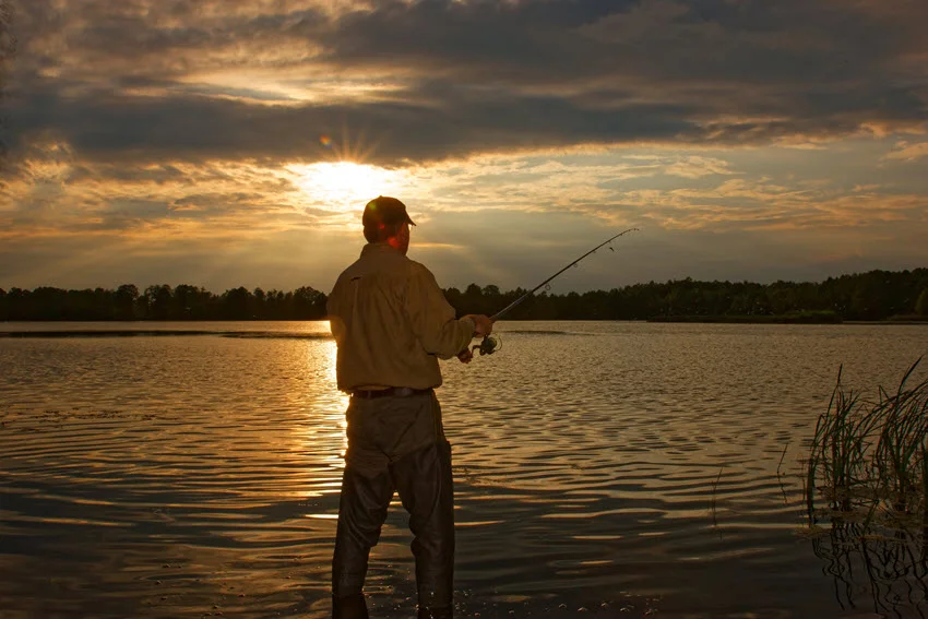 Angler in wade boots, stand in lake shore and fish, during sunset.