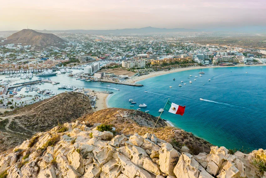 A Mexican flag overlooks Cabo San Lucas, where boats fill the marina and bay beside a sandy beach and coastal buildings.