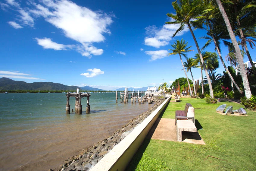 Palm trees and benches line the Cairns waterfront in Queensland, Australia, with wooden pilings in the water and mountains beyond.