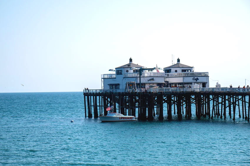 White buildings stand at the end of a wooden California pier, with a lifeguard boat alongside in the blue Pacific water.