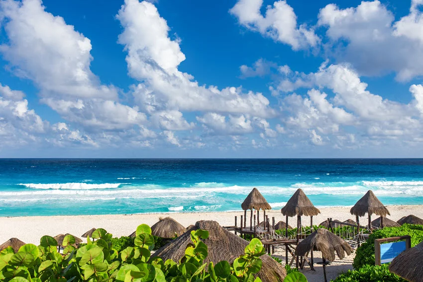 Thatched beach umbrellas line Playa Delfines in Cancún, Mexico, overlooking white sand and turquoise Caribbean surf beneath scattered clouds.