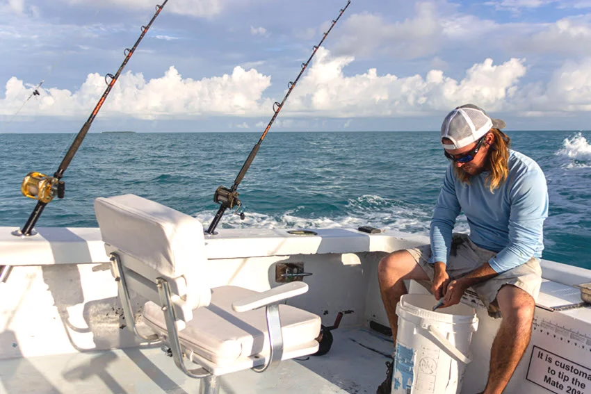 A crew member prepares bait aboard a Key West charter boat, with fishing rods mounted along the stern and ocean wake behind.