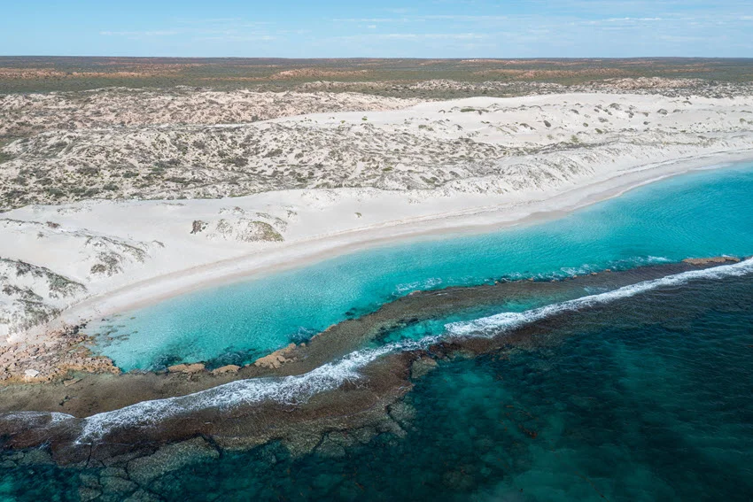 White sand and coastal dunes border turquoise shallows near Exmouth, Western Australia, while waves break over a rocky offshore ledge.