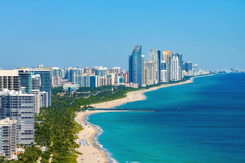 Beachfront high-rises line the Miami-area coastline, with sandy beaches and short rock jetties bordering turquoise Atlantic water beneath a blue sky.