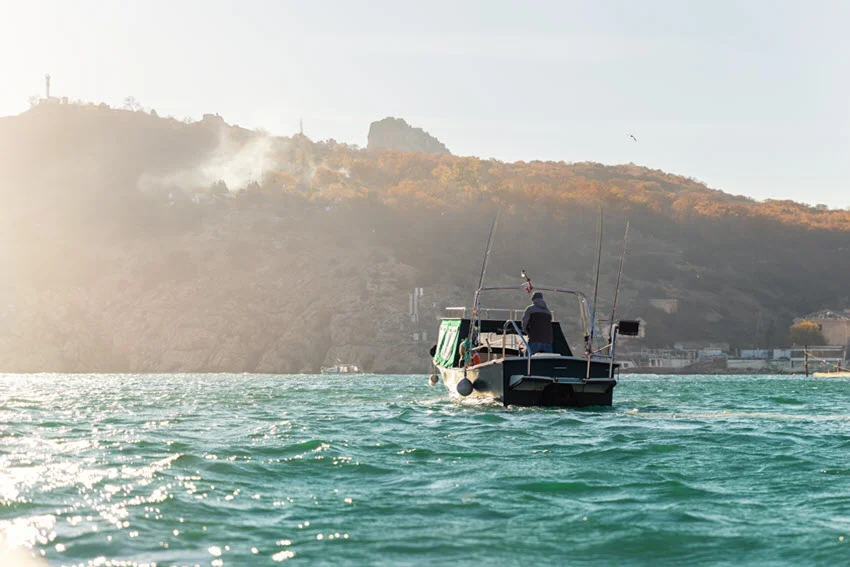 An angler rides aboard a small fishing boat with upright rods as morning light illuminates the rocky shoreline of a harbor.
