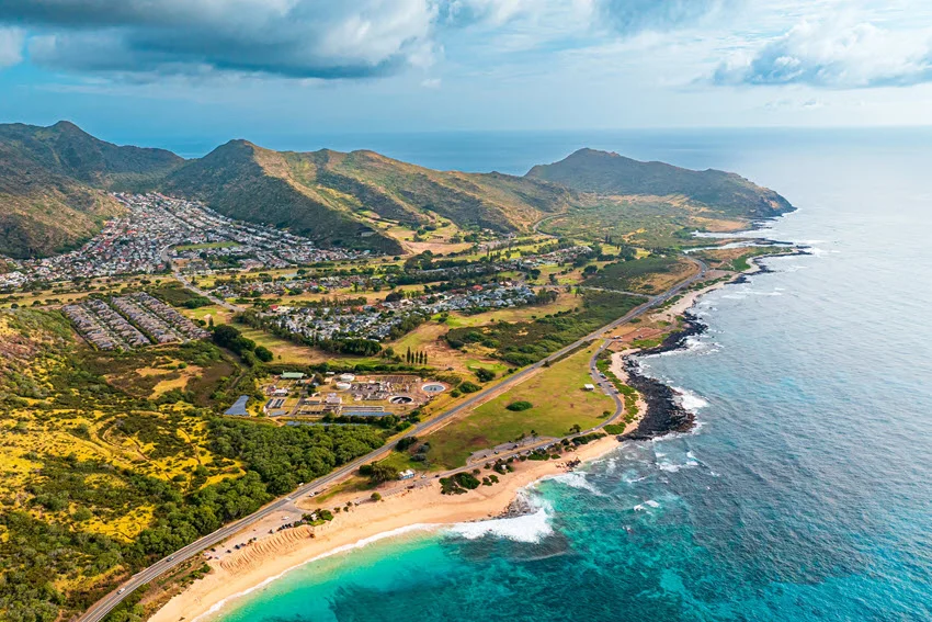 A coastal road follows Oahu's shoreline in Hawaii, winding between steep green mountains, sandy beaches, and waves breaking over coastal rocks.
