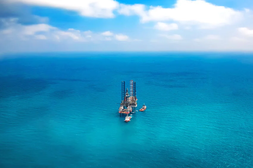 An offshore oil rig stands in blue ocean water with support vessels alongside, viewed from above beneath scattered clouds.