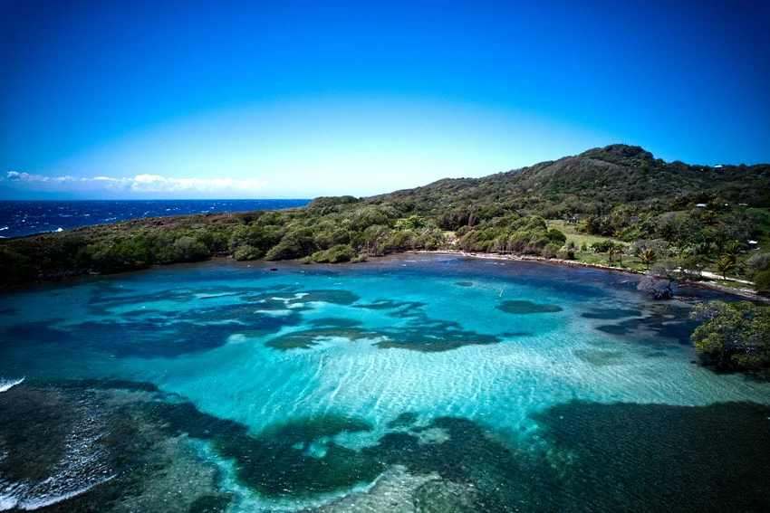 Clear turquoise water reveals rippled sand and dark underwater patches in a cove on Roatán, Honduras, bordered by green hills.