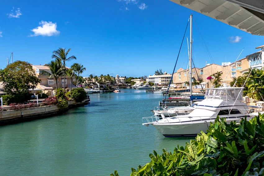 Sailboats and motor yachts sit in a sheltered marina in St. Peter, Barbados, surrounded by waterfront homes and palm trees.