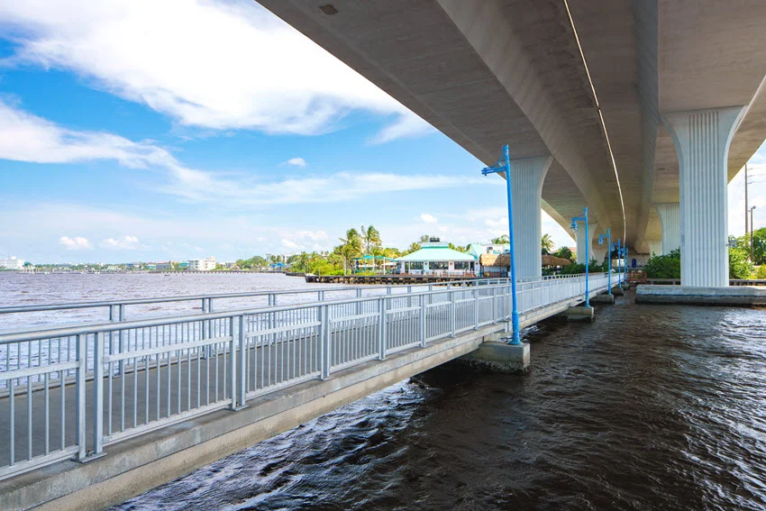 The Riverwalk in Stuart, Florida, passes beneath a concrete bridge, with white railings and blue lamps beside the waterfront.