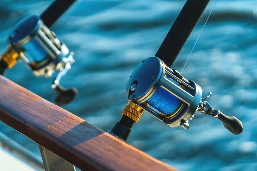 Focused view of two conventional reels spooled with blue line and mounted on rods beside a wooden boat rail.