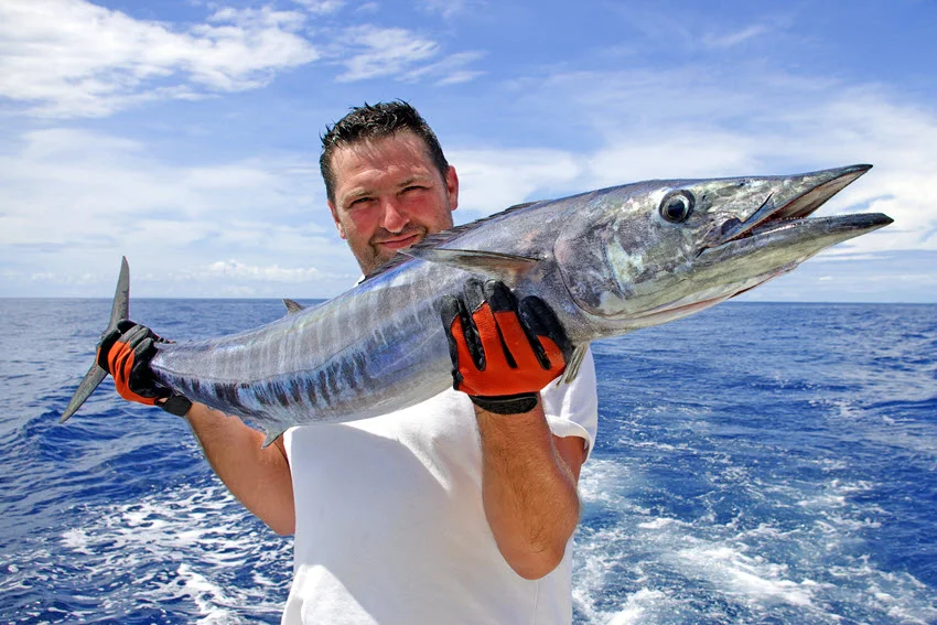 A happy angler holding a large Wahoo aboard a fishing boat in deep offshore waters.