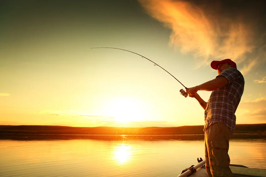 Side view shot of a man casting his fishing rod against a golden sunset, with warm light illuminating his silhouette and the water below.
