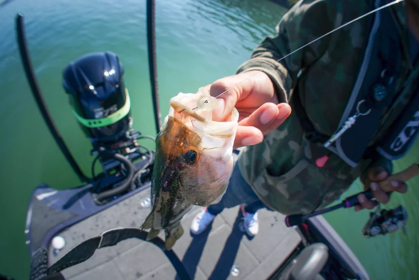 An angler holds a Bass beside a boat motor, with a small billed crankbait hooked near its mouth and fishing line extending upward.