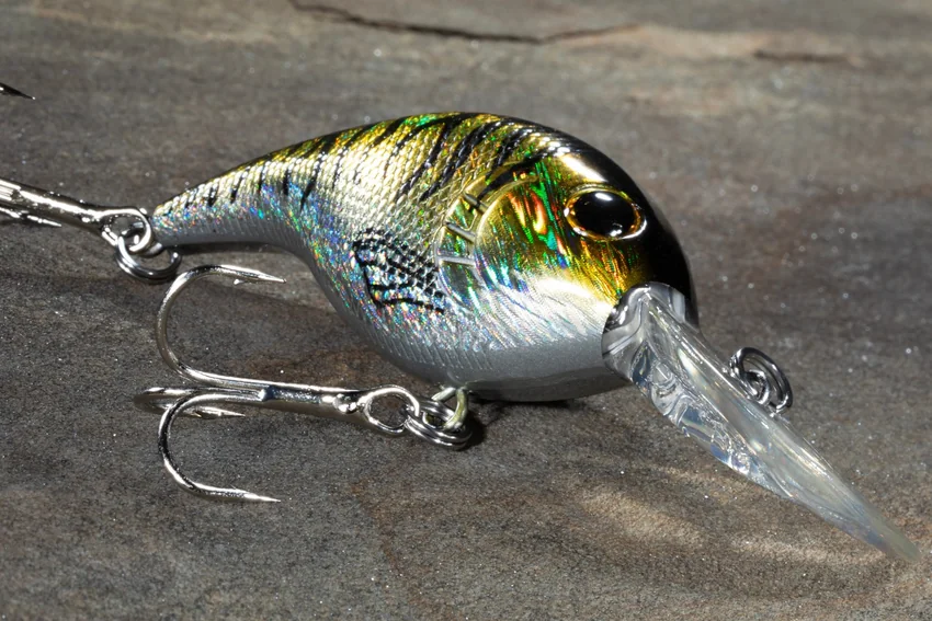 A rounded crankbait with a reflective green and silver body, clear diving bill, and treble hooks rests on a textured gray surface.