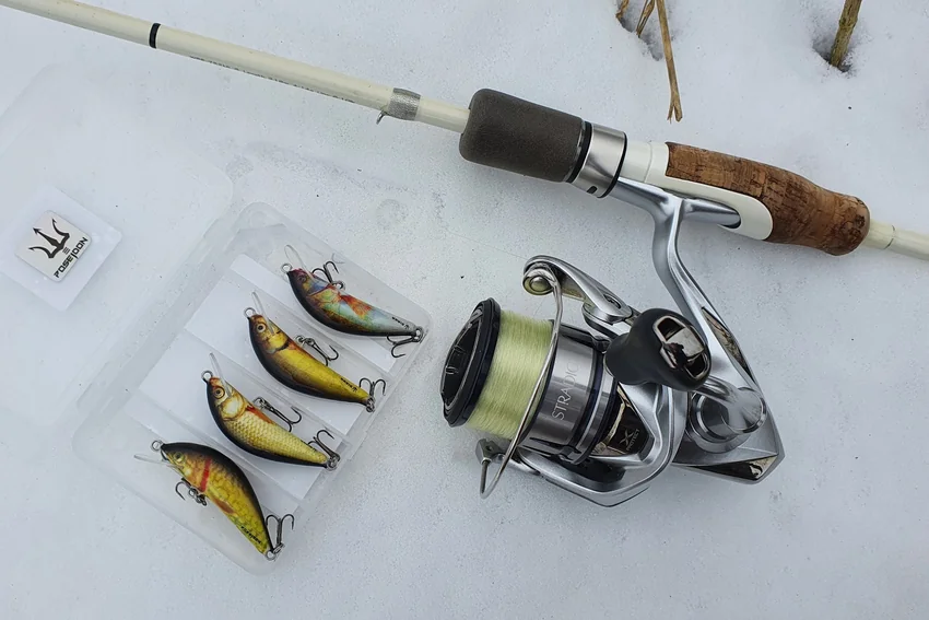 A spinning rod and reel lie on snow beside an open tackle box containing four hard-bodied lures with different colors and body profiles.