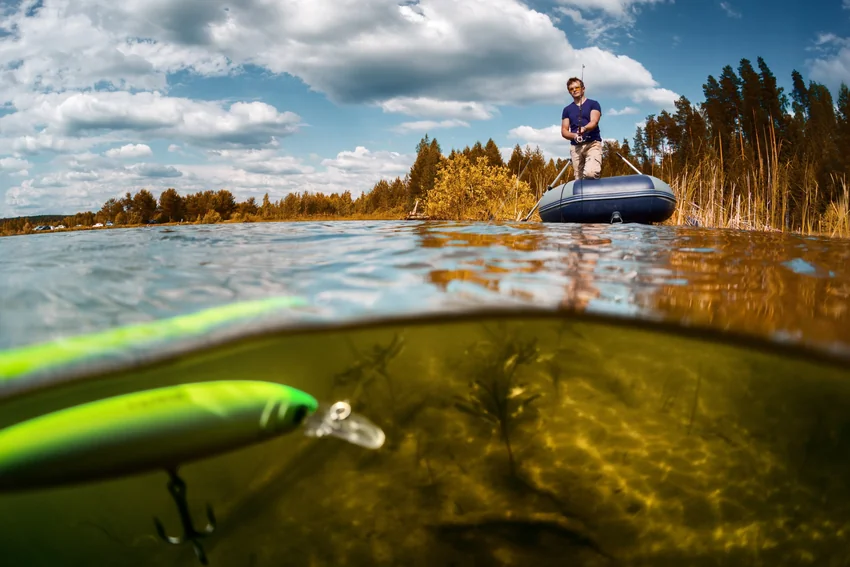 A green crankbait appears beneath the waterline in the foreground while an angler fishes from an inflatable boat beside reeds and autumn trees.