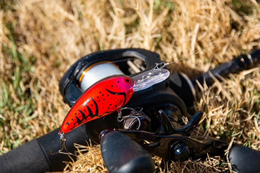 A red crankbait with black markings, a clear diving bill, and treble hooks rests across a baitcasting reel on dry grass.