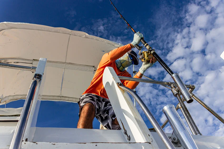 A bottom-up view of an angler in full protective gear, wearing sunglasses and a cap, securing a fishing rod into a holder on a fishing charter boat. The bright blue sky with scattered white clouds sets the scene on a sunny day.