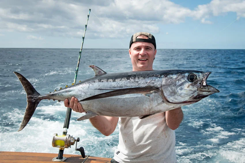 An angler smiling while posing with a Tuna trophy held in both hands on the wooden deck of the charter boat, with open water stretching out in the background.