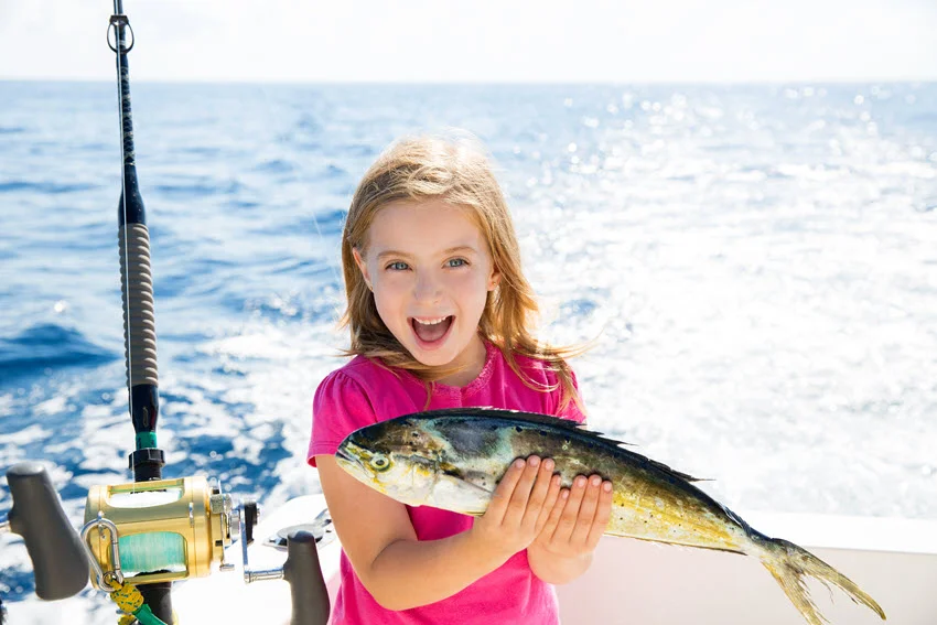 A happy blonde girl posing on a boat deck while holding a Mahi Mahi fish, with the open ocean in the background and a fishing rod beside her.